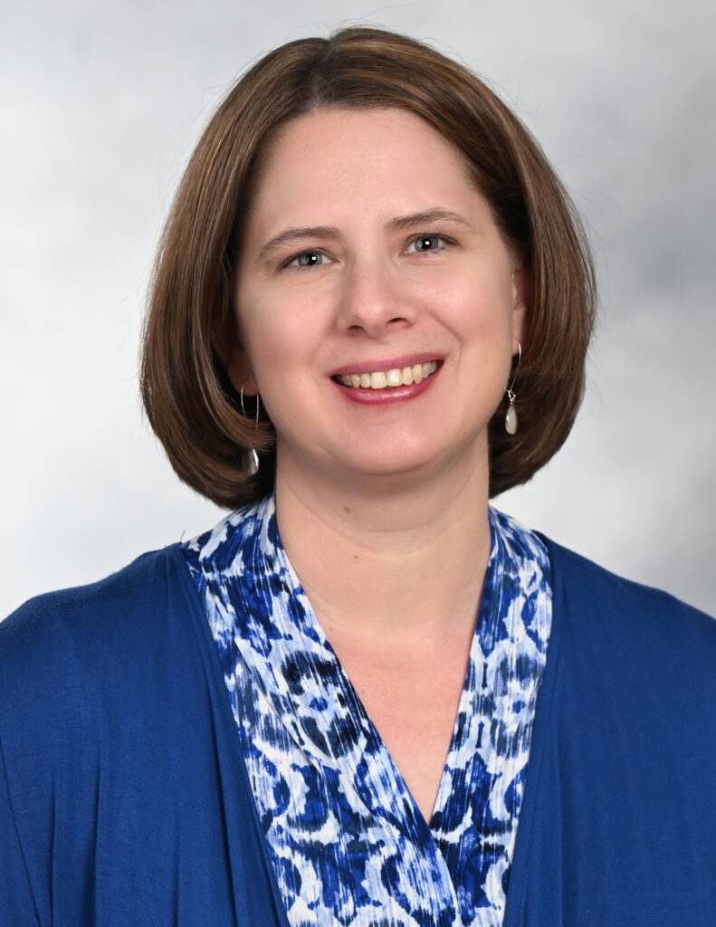 White female in her late 40s with shorter brown hair wearing a blue cardigan over a blue patterned shell.