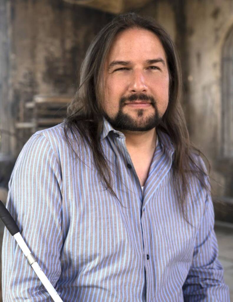 Headshot of Joe with white cane. Joe is wearing a button-up shirt, is in focus with the background blurred behind him.