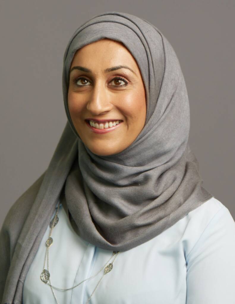 Smiling pretty woman wearing a grey head scarf and light blue shirt and a silver necklace.