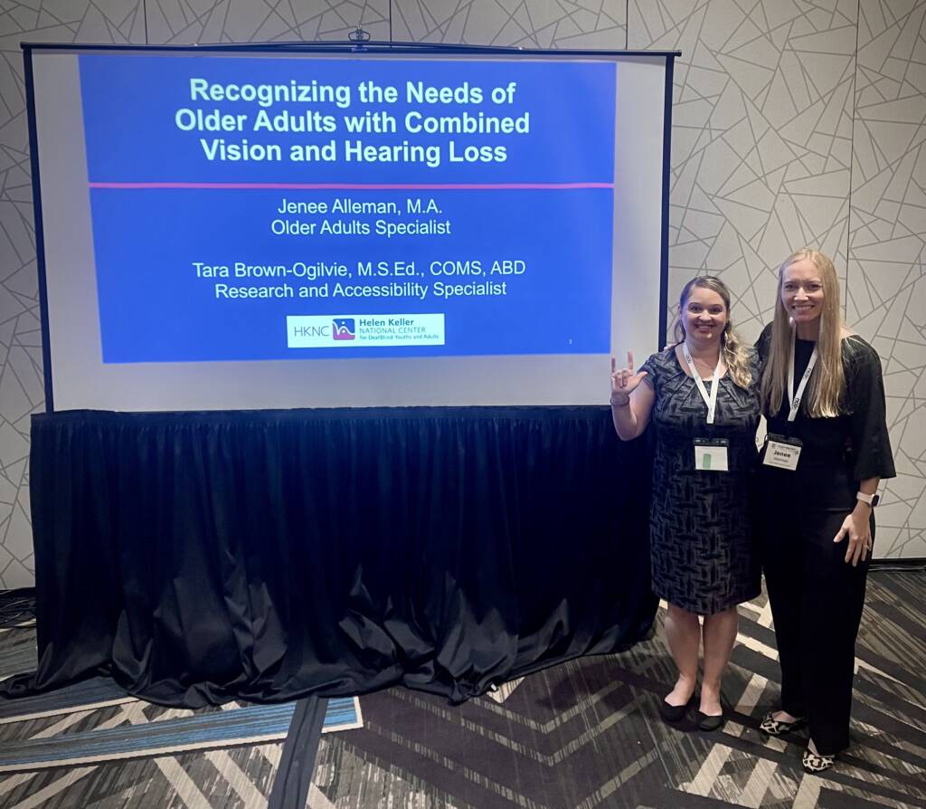 HKNC staff Tara Brown-Ogilvie and Jenee Alleman stand to the right of their presentation entitled, "Recognizing the Needs of Older Adults with Combined Vision and Hearing Loss." They are wearing business clothing with name badges in a conference setting.