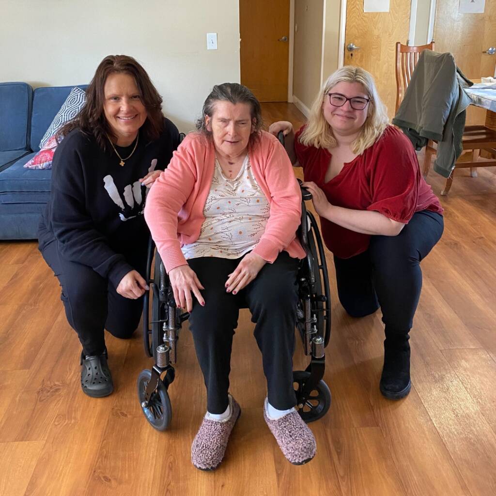3 smiling women. One of the women sits in a wheelchair and the other 2 women kneel on either side of her.