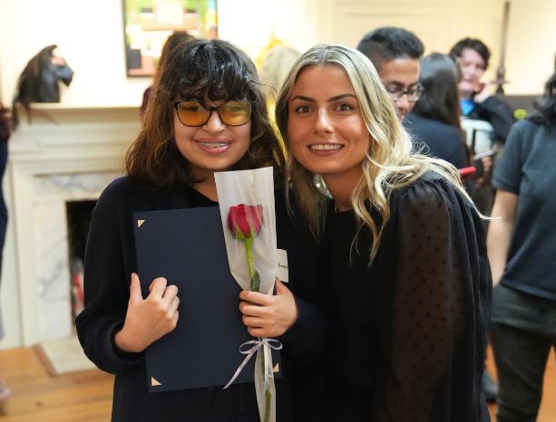 A young woman stands next to her art instructor in an art gallery and holds a red rose.