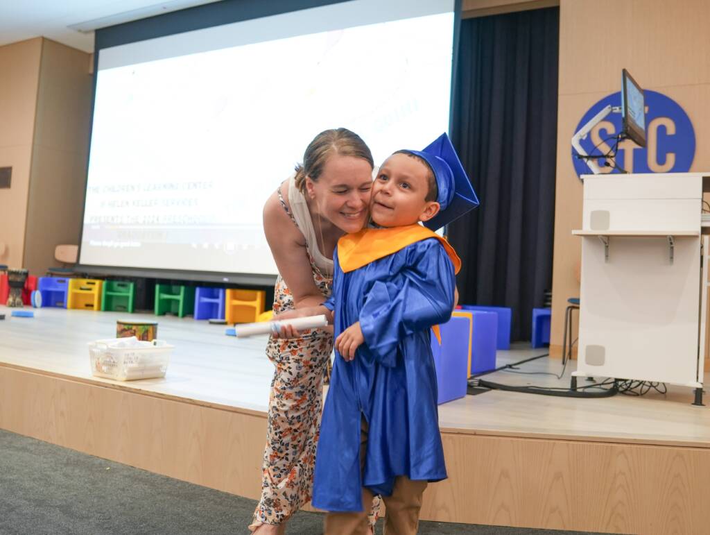 A smiling woman hugs a preschool boy who wears a blue and gold graduation cap and gown and holds a rolled-up diploma.