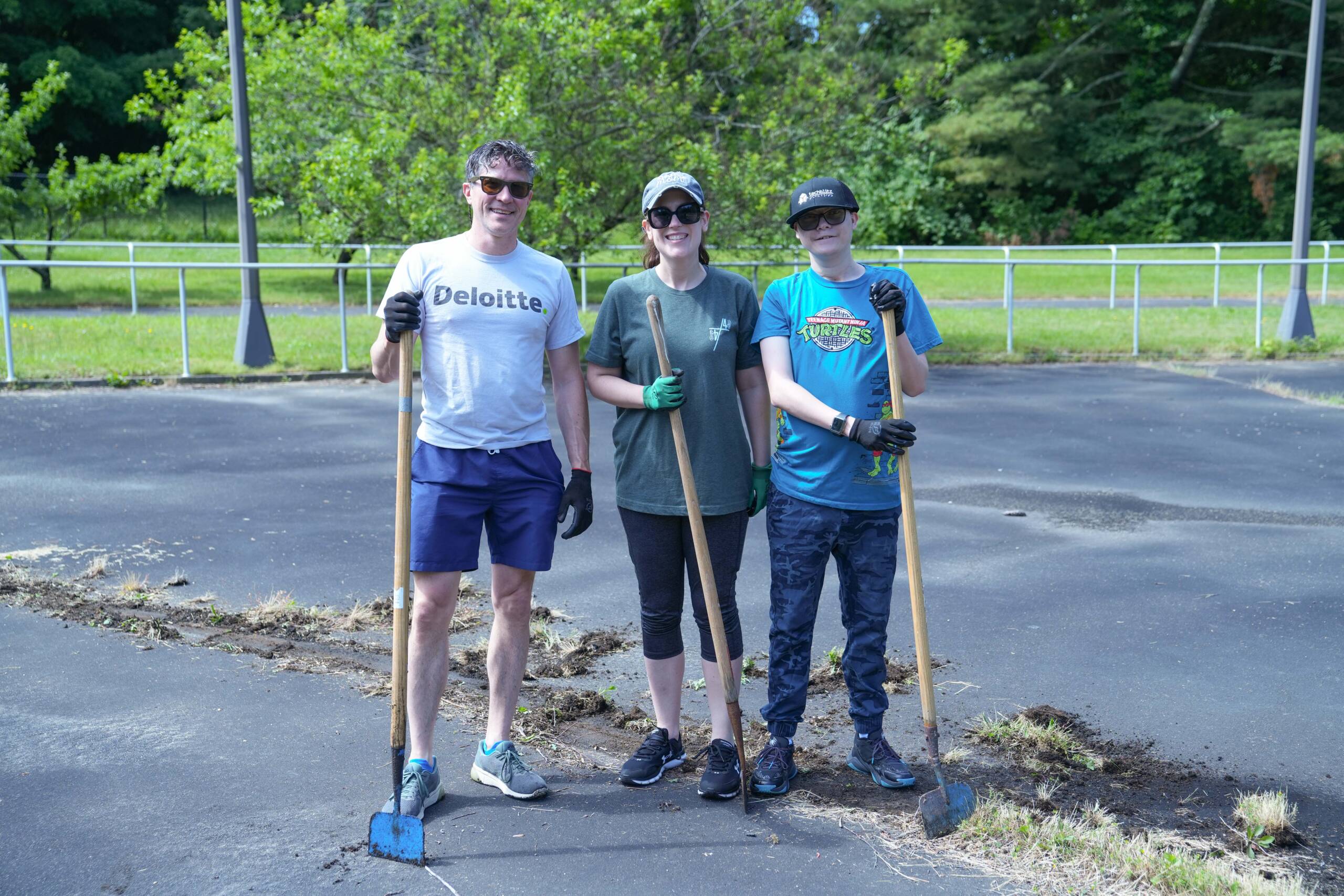 A Deloitte volunteer, a HKS female staff member, and a male participant smiling while holding gardening tools. All three of them are wearing gardening gloves and black sunglasses.