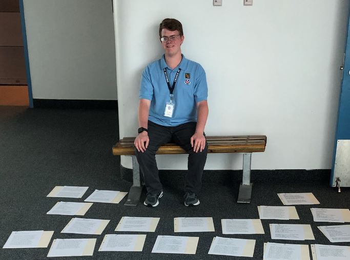 A man sits on a bench in front of dozens of pieces of paper on the floor that show braille descriptions and labels.