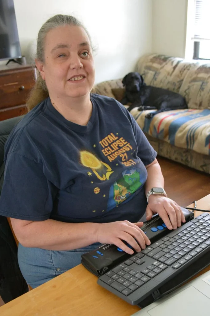 A woman smiling and using a braille display. Her dog guide is in the background.