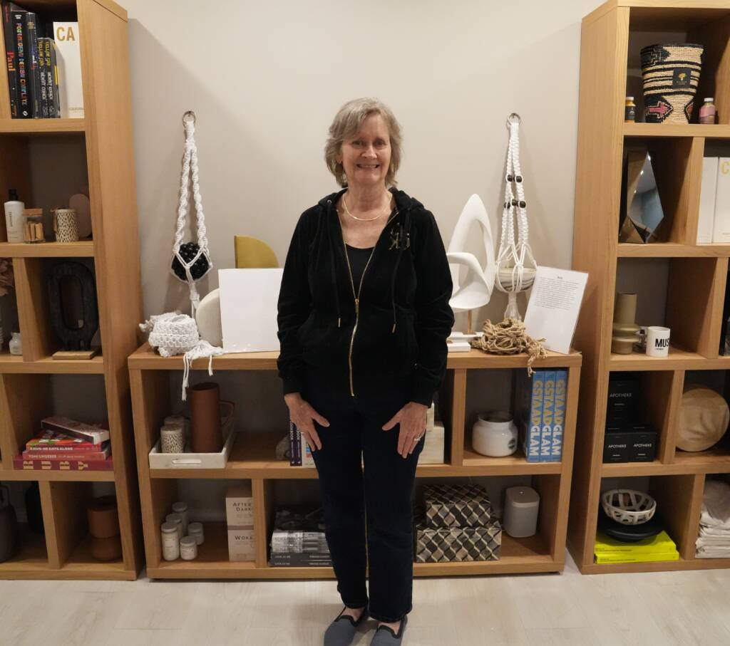 An older woman stands in front of 2 macrame hangers she made.