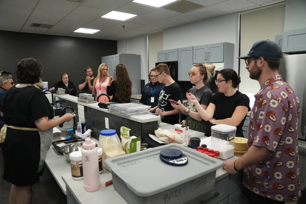 A group of people stand around a kitchen. Bagel-making supplies are on the countertops.