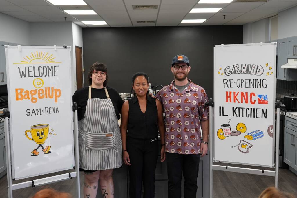 3 people smile in between 2 large white boards that say "Welcome BagelUp Sam & Reva" and "Grand Re-Opening HKNC IL Kitchen" on them.