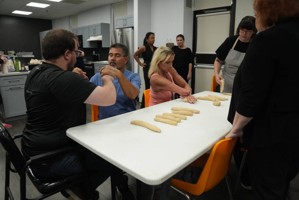 Two people communicating in tactile sign language next to a woman kneading dough with one hand.