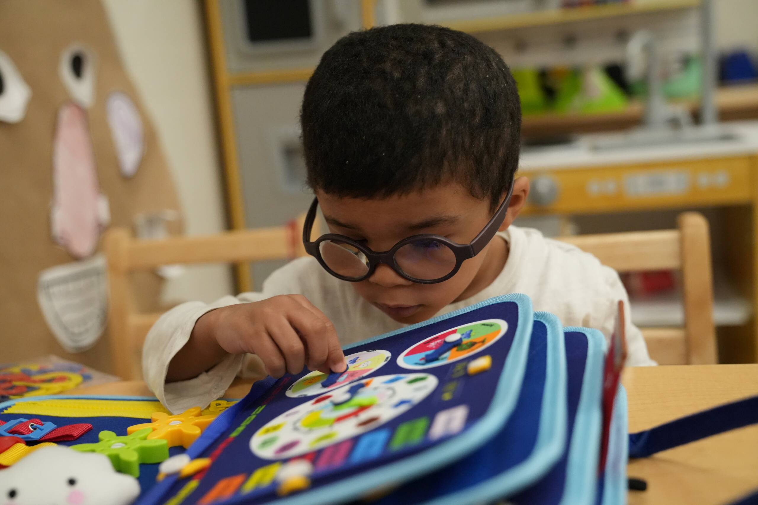 Upclose image of a pre-school boy wearing glasses interacting with a tactile learning book in HKSB's Children's Learning Center. 