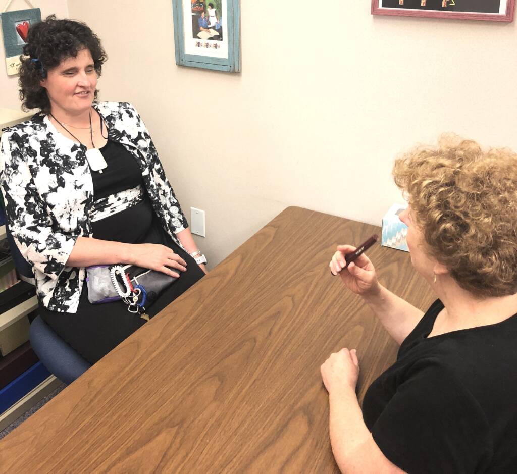 Two women are sitting across from each other at a table. One woman is wearing an FM Loop around her neck while the other woman speaks into the FM Microphone.