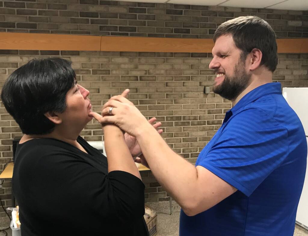 A man and woman are standing in front of each other. The woman is signing while the man tactually has his hands on her hands. The man is laughing.