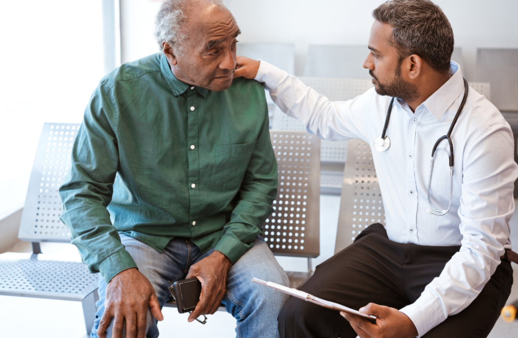 An older man is sitting to the left of a doctor. The doctor has his hand on the mans shoulder and is speaking to him.