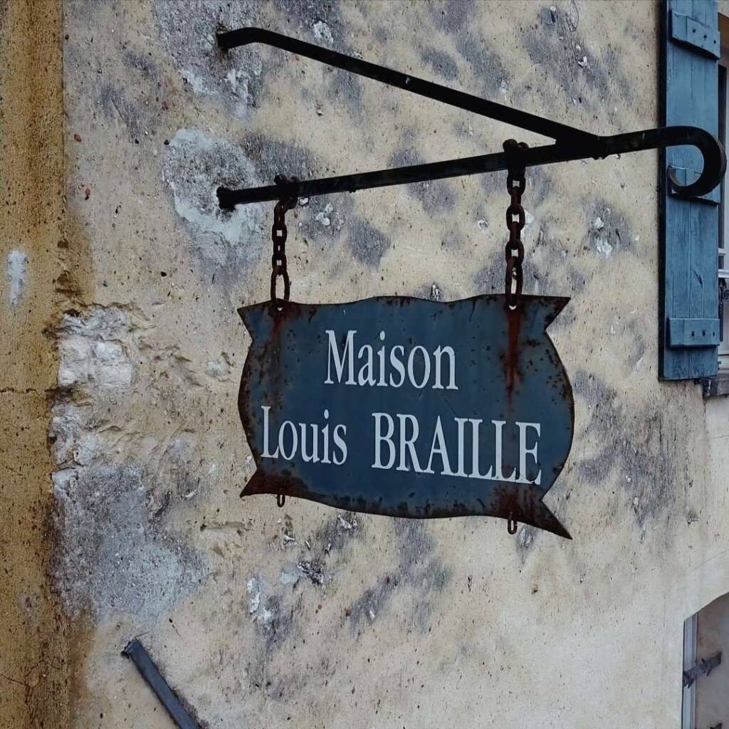 A weathered hanging sign mounted on an exterior wall of the Louis Braille Museum reads "Maison Louis BRAILLE" in white text on a dark blue background.