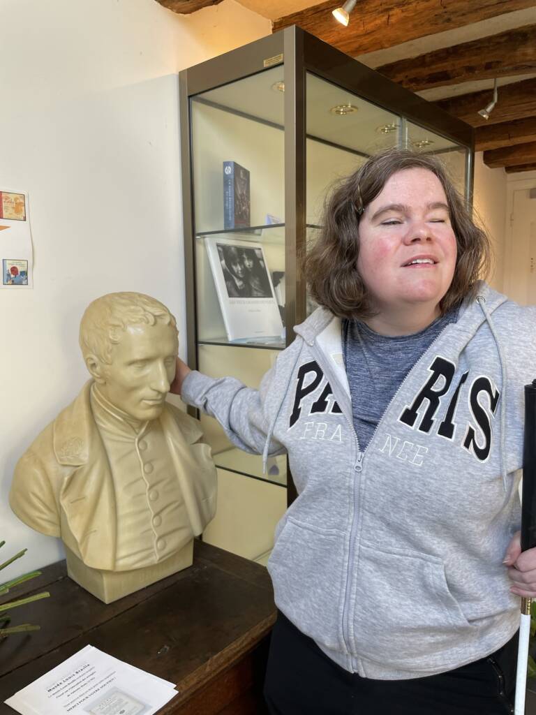 Megan standing in the Louis Braille Museum while touching a statue bust of Louis Braille.