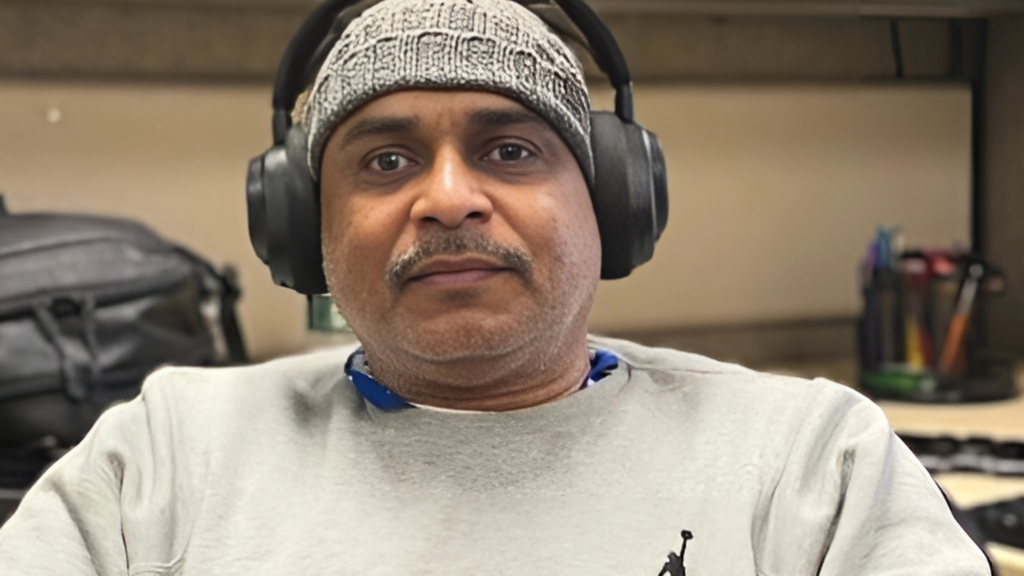 A man waering a hat and headphones, wits at his desk smiling at the camera.