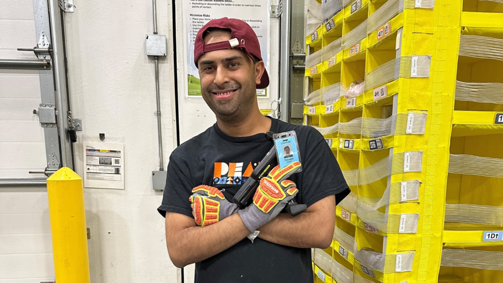 Faruqee stands in an Amazon warehouse smiling while holding his employee badge and a product scanning device.