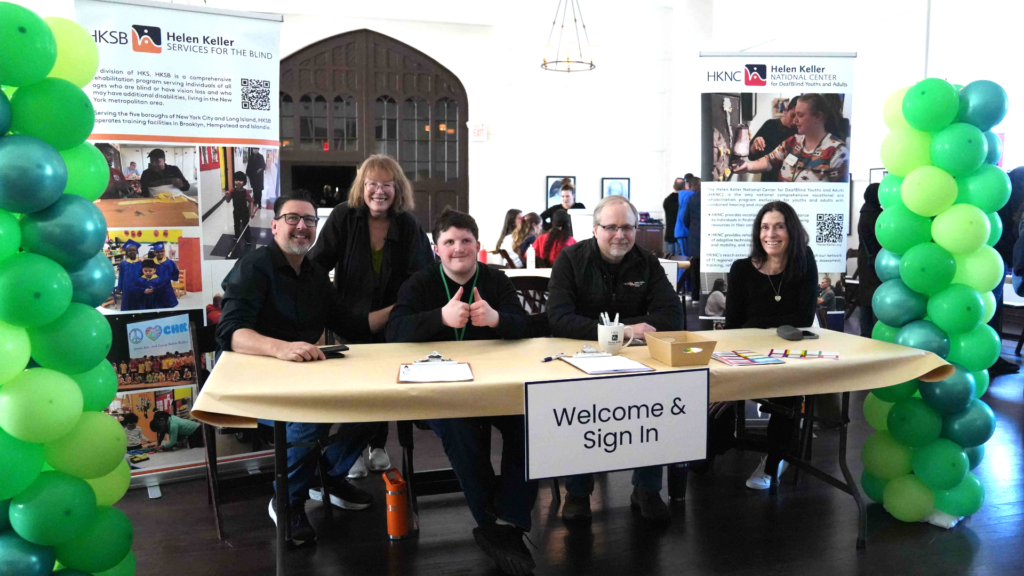 Three adults HKS staff members and a young adult sitting at a welcome table with one adult staff member standing with them. The table has balloon displays on each side of it as well as two standing banners behind it.