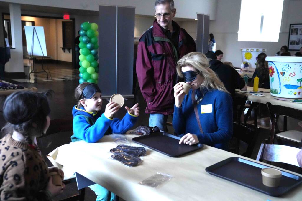 An adult female wearing a blind fold smells a small container while a young girl looks on smiling.