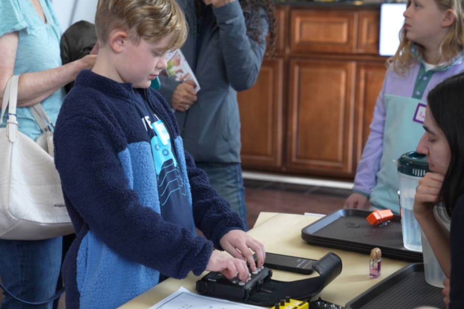 A child touches a braille display while adults look on. 