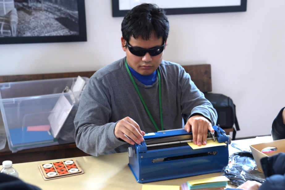 A young adult is brailling a name card on a Perkins Brailler.