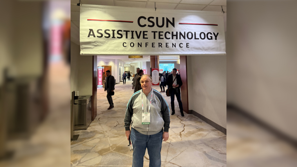 1. Kevin standing beneath the CSUN Assistive Technology Conference banner in the main conference hall.